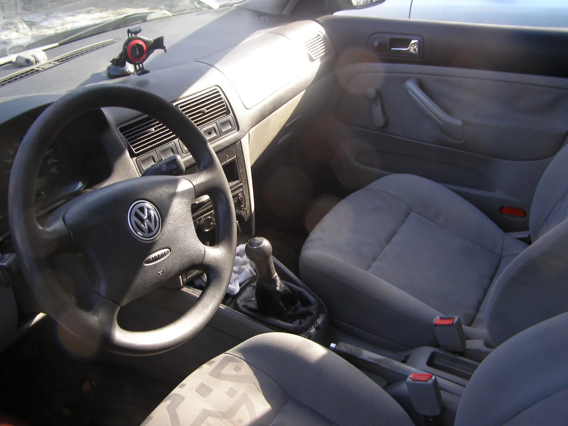 Wnętrze samochodu marki Volkswagen z widocznym kierownicą z logo VW, szarymi siedzeniami, dźwignią zmiany biegów, deska rozdzielcza z wentylacjami i przyciskami, uchwytem na drzwiach oraz uchwytem na przednią szybę z czerwonym elementem. Widoczne są również czerwone przyciski zapięć pasów bezpieczeństwa i fragment zewnętrzny przez przednią szybę.