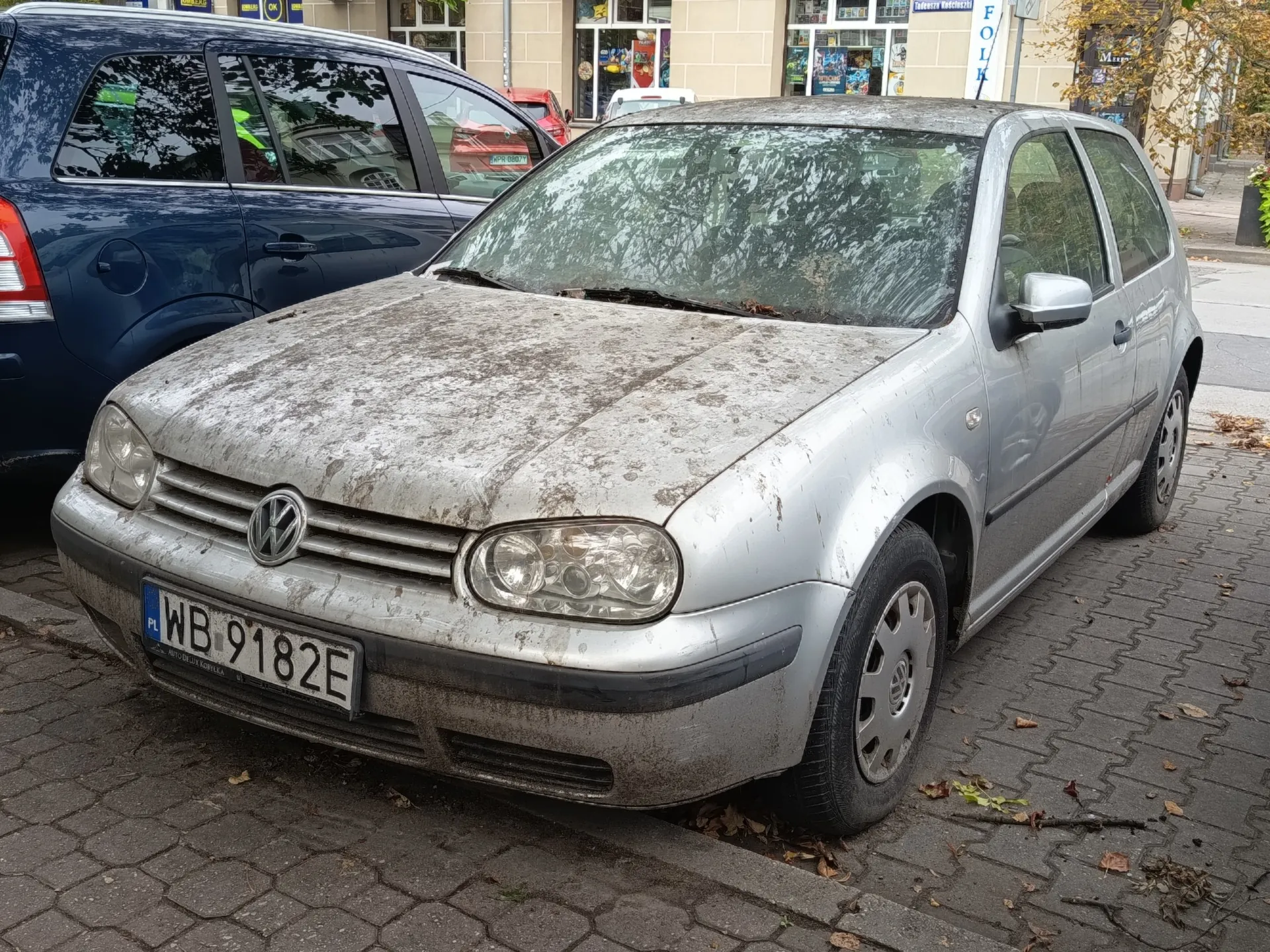 Srebrny samochód osobowy marki Volkswagen Golf zaparkowany na kostce brukowej. Pojazd jest bardzo brudny, pokryty warstwą kurzu i błota, szczególnie na masce i przednim zderzaku. Samochód posiada polską tablicę rejestracyjną o numerze WB 9182E. Obok niego, po lewej stronie, stoi ciemnoniebieski samochód typu kombi.