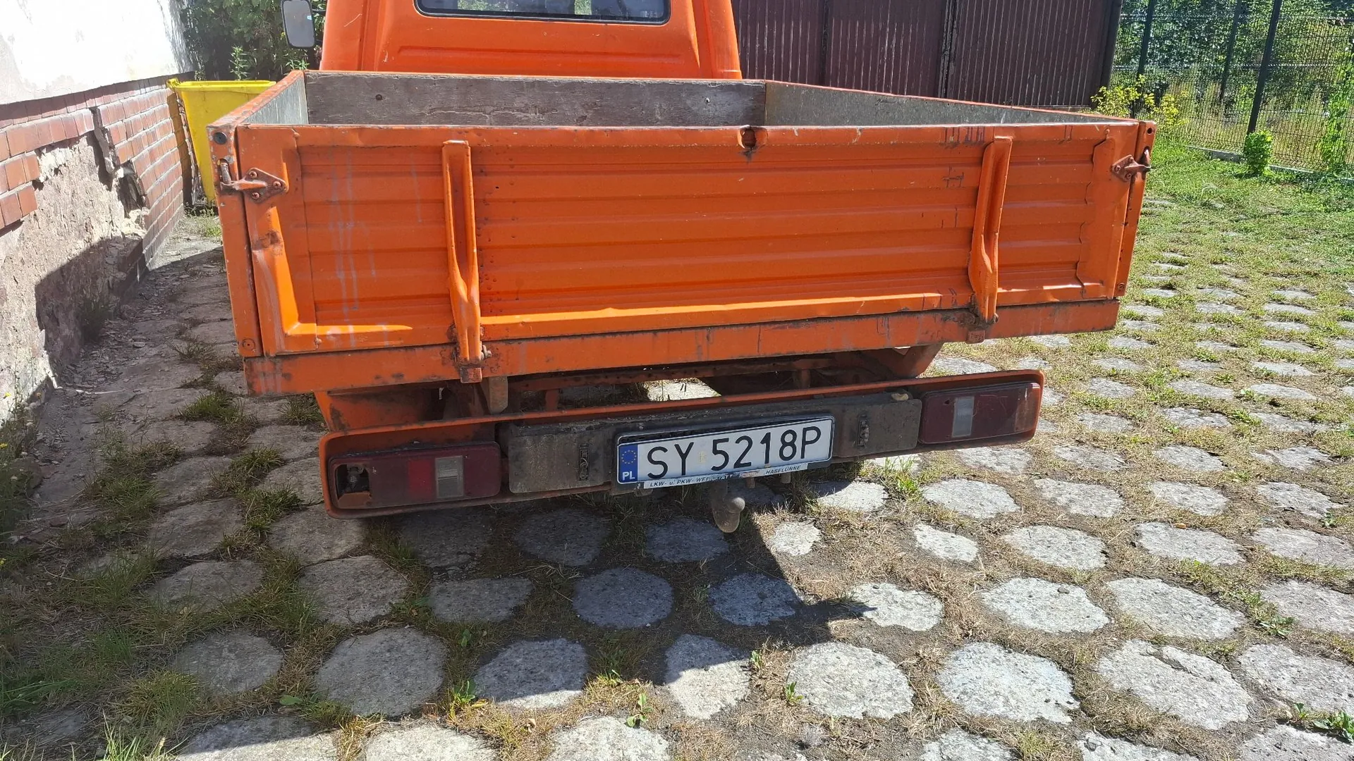 Tylny widok pomarańczowej ciężarówki z otwartą skrzynią ładunkową, zaparkowanej na podjeździe z kostki brukowej i trawy. Pojazd posiada tablicę rejestracyjną o numerze SY 5218P.