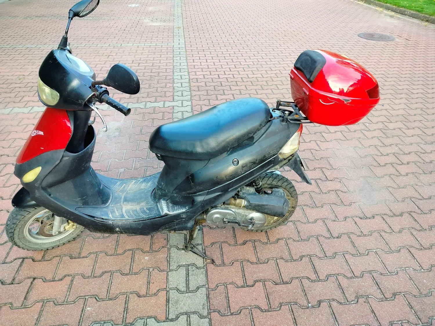 Czarny skuter z czerwonymi elementami nad przednim kołem oraz czerwonym kufrem zamontowanym z tyłu. Pojazd stoi na kostce brukowej w kolorze czerwonym i szarym.
