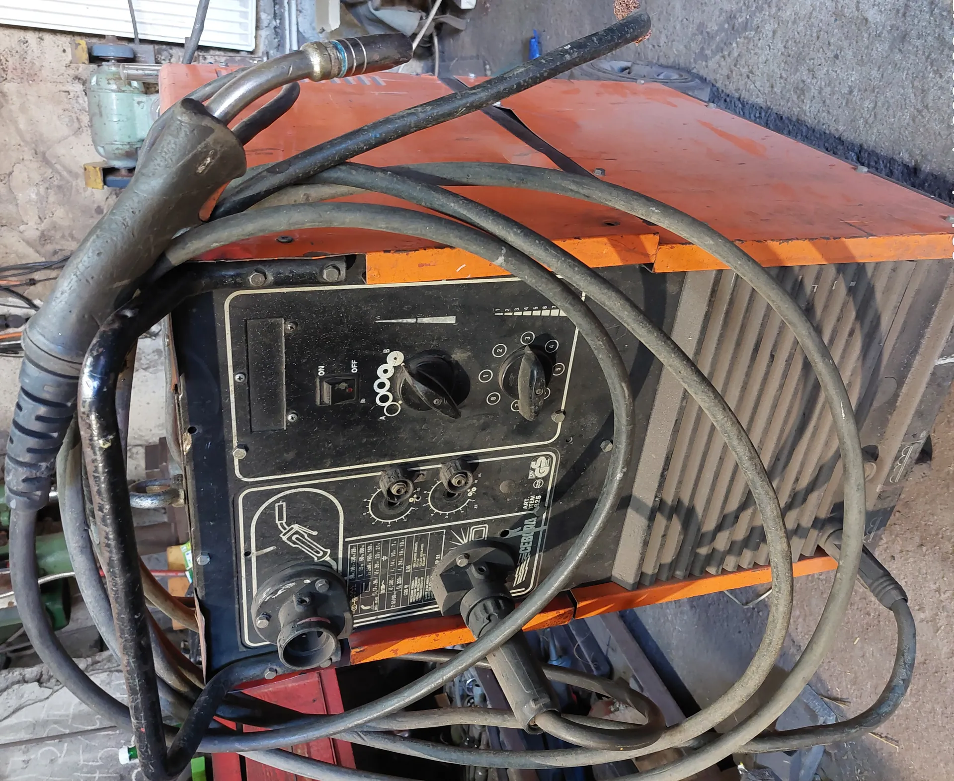 Stacja spawalnicza marki CEBORA, model ART. 325, o pomarańczowo-czarnym kolorze, z wieloma przewodami owiniętymi wokół urządzenia. Panel sterowania zawiera przełącznik ON/OFF, pokrętła, wskaźniki oraz schematyczny rysunek pistoletu spawalniczego. W tle widoczne są betonowe ściany warsztatu i inne urządzenia.