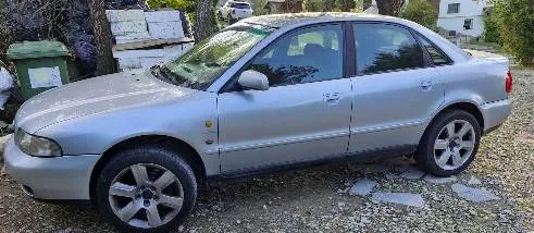 Srebrny samochód osobowy typu sedan zaparkowany na żwirowym podjeździe. Pojazd jest pokazany z profilu od strony kierowcy. W tle widoczny jest zielony kosz na śmieci, drzewa oraz fragment białego budynku.