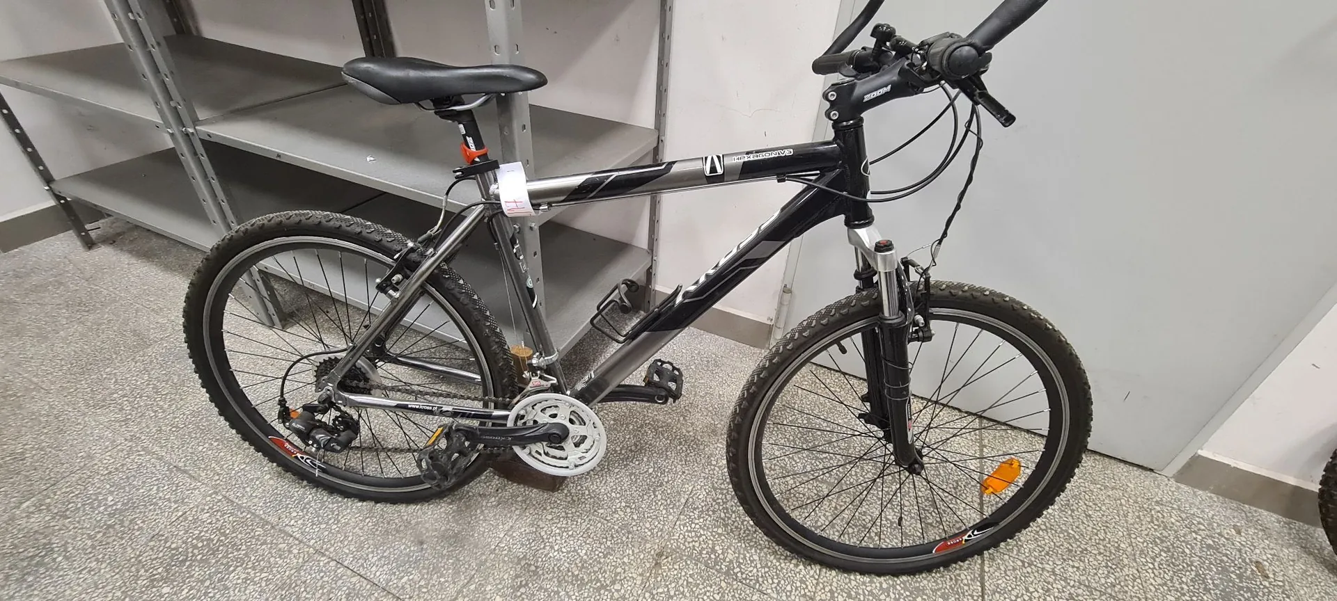 Czarny rower górski z białymi i szarymi akcentami na ramie, zaparkowany w pomieszczeniu na szarej, chropowatej podłodze. Rower posiada amortyzator przedni, czarne opony z bieżnikiem, czarne siodełko oraz srebrną korbę. W tle widoczne są metalowe regały magazynowe.