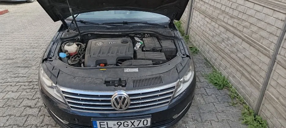 Widok z przodu na ciemny samochód marki Volkswagen z otwartą maską, odsłaniającą silnik z napisem TDI. Samochód posiada polską tablicę rejestracyjną o numerze EL 9GX70. Pojazd stoi na kostce brukowej obok jasnego, betonowego ogrodzenia.