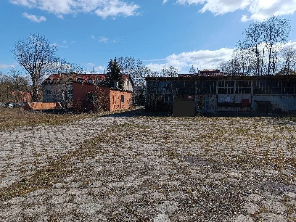 Zdjęcie przedstawiające zniszczony, betonowy plac z wyrastającą trawą. W tle znajdują się dwa budynki: po lewej czerwony, dwupiętrowy budynek mieszkalny z czerwonym dachem, a po prawej niski, szary budynek przemysłowy lub magazynowy z licznymi oknami. Nad budynkami widoczne jest jasnoniebieskie niebo z białymi chmurami oraz kilka bezlistnych drzew.