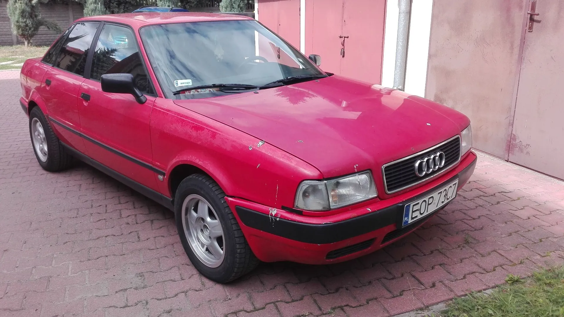 Czerwony samochód marki Audi zaparkowany na kostce brukowej przed różową ścianą budynku. Pojazd ma widoczną tablicę rejestracyjną o numerze EOP 73C7. Na masce i przednim zderzaku widoczne są drobne uszkodzenia lakieru.