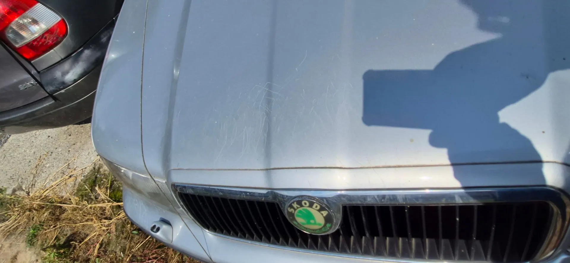Zbliżenie na maskę i grill srebrnego samochodu marki Skoda. Na powierzchni maski widoczne są liczne zarysowania. W prawym górnym rogu widoczny jest cień osoby robiącej zdjęcie. Po lewej stronie fragment ciemnego samochodu z widoczną tylną lampą.