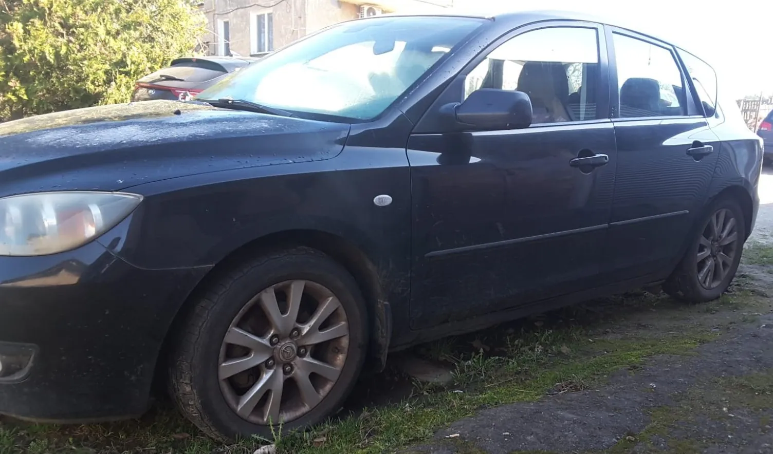 Czarny samochód osobowy zaparkowany na trawie i żwirze. Pojazd jest zabrudzony, widoczne są felgi aluminiowe. W tle drugi samochód oraz budynek z oknami i zieleń.