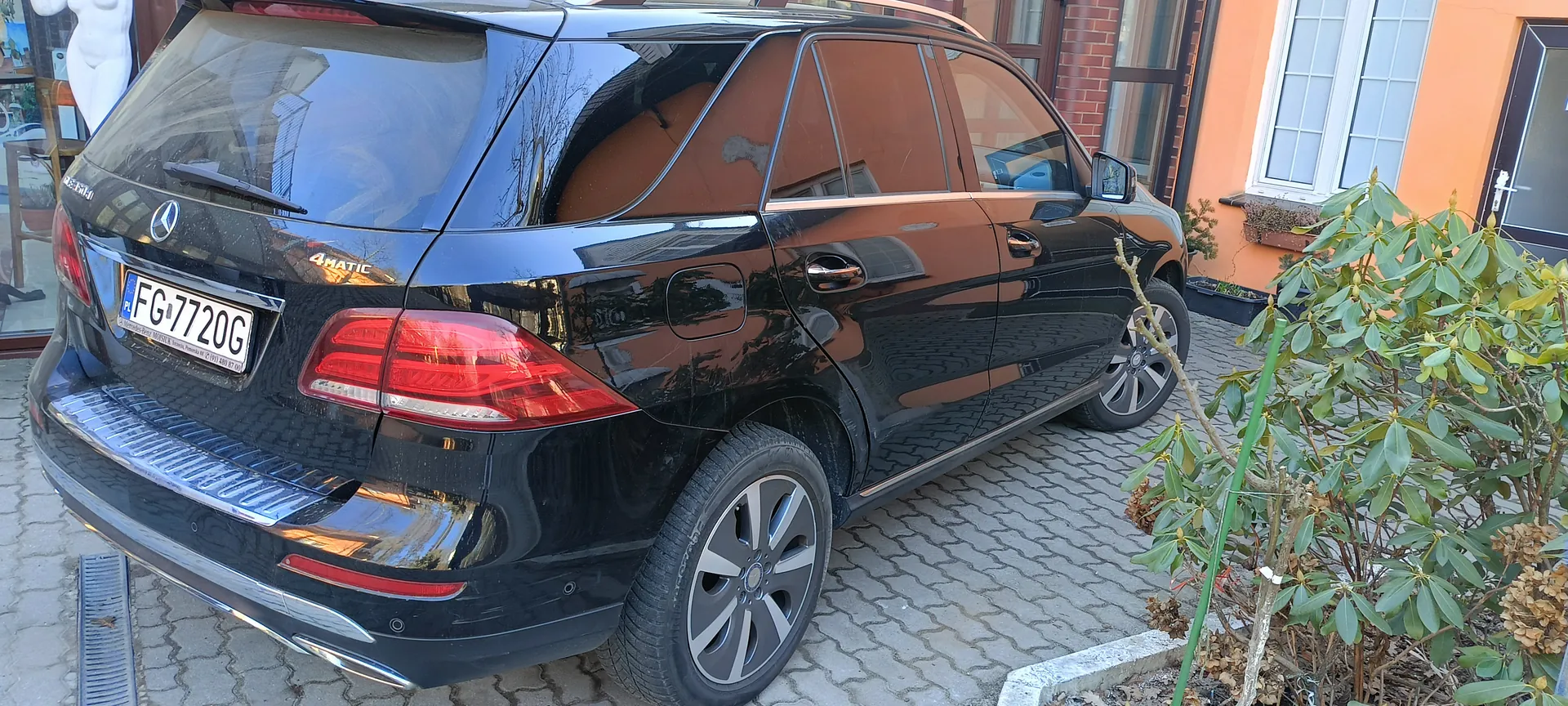 Czarny samochód Mercedes-Benz typu SUV zaparkowany na brukowanej powierzchni. Widoczna tylna część pojazdu z tablicą rejestracyjną PL FG-7720G oraz napisem 4MATIC na bagażniku. Po prawej stronie obrazu krzew z zielonymi liśćmi, w tle pomarańczowy budynek z białymi oknami i ceglaną ścianą. Odbicia na błyszczącej karoserii samochodu ukazują otoczenie.