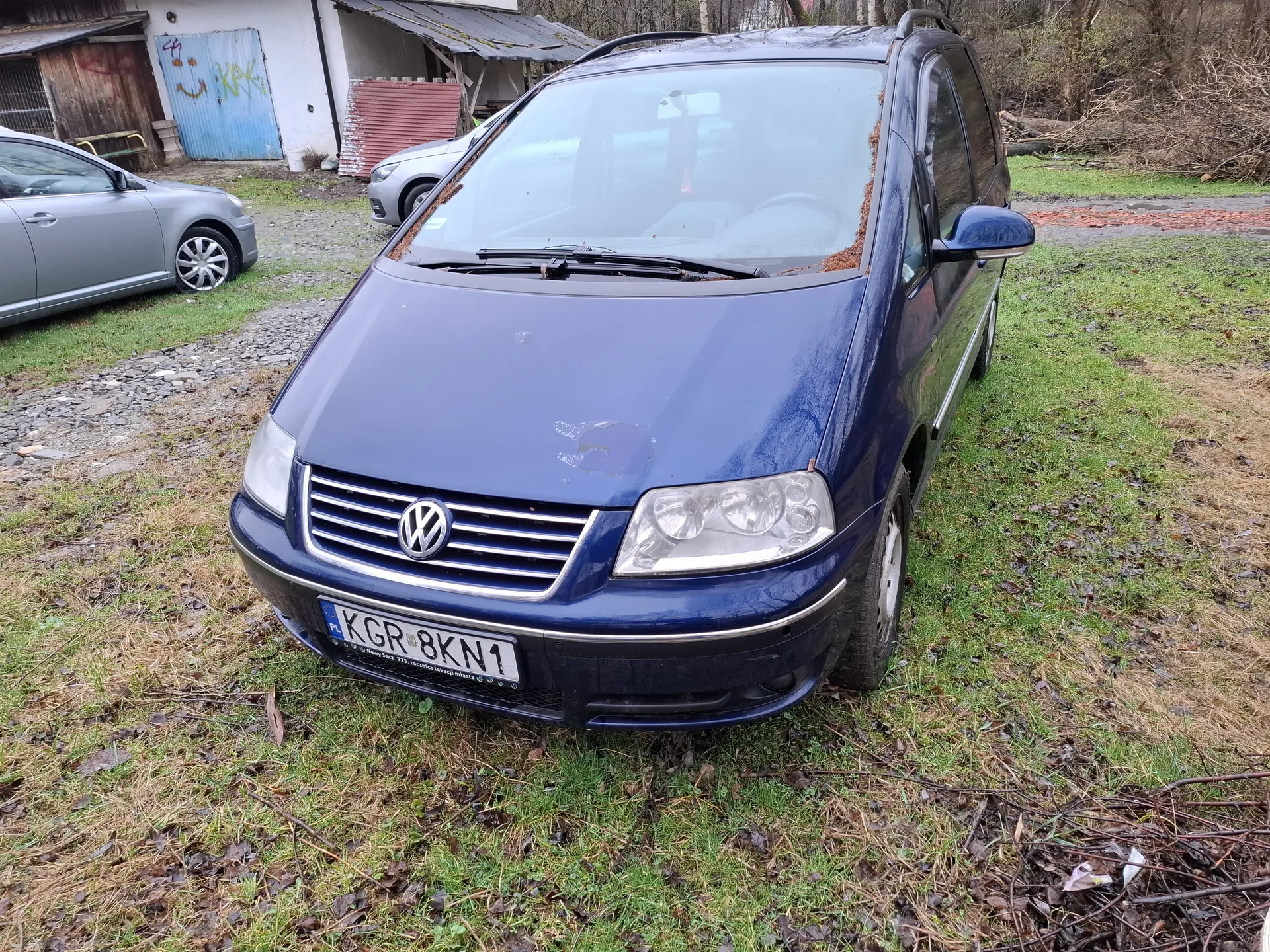 Niebieski samochód osobowy marki Volkswagen z tablicą rejestracyjną KGR 8KN1, zaparkowany na trawie z suchymi liśćmi. W tle widoczne są dwa inne pojazdy: srebrny samochód po lewej stronie oraz częściowo widoczny pojazd za niebieskim Volkswagensem. W tle również budynek z niebieską bramą pokrytą graffiti oraz czerwonym elementem konstrukcyjnym. Trawa jest częściowo wyschnięta, a na ziemi leżą gałęzie i liście.