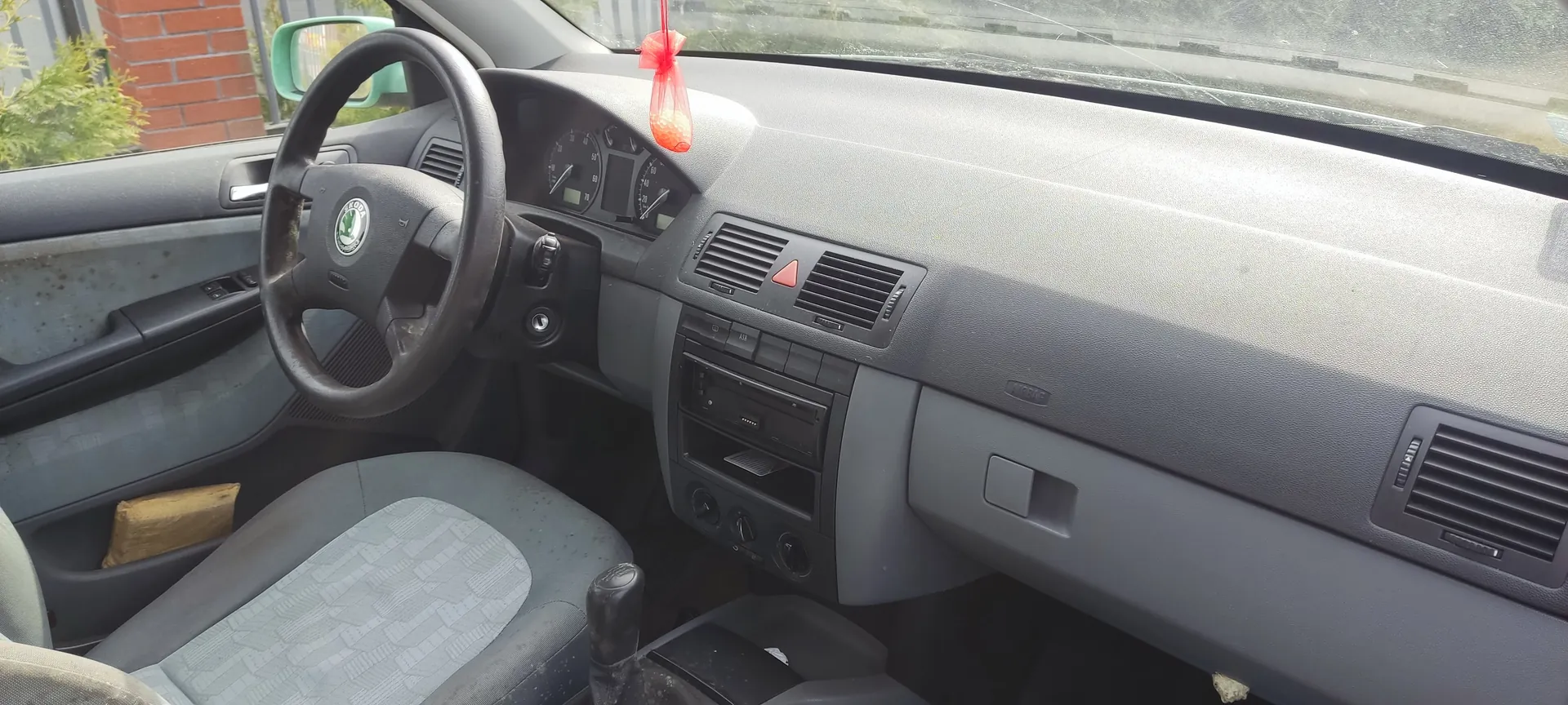 Wnętrze samochodu z kierownicą marki Škoda, szarą deską rozdzielczą z widocznym napisem "AIRBAG", czerwonym zapachem wiszącym na lusterku wstecznym, szarymi siedzeniami z geometrycznym wzorem, dźwignią zmiany biegów, wentylacjami i radioodtwarzaczem. Po lewej stronie widoczna ceglana ściana i zieleń za oknem. W kieszeni drzwi znajduje się żółty przedmiot. Przyrządówka z licznikami prędkości i obrotomierzem.