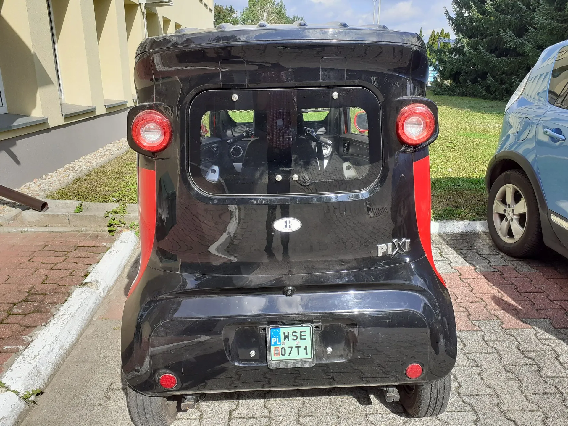 Tylny widok małego, czarnego samochodu elektrycznego marki Piaggio z czerwonymi akcentami po bokach. Pojazd posiada okno z tyłu, dwa okrągłe czerwone światła stop oraz tablicę rejestracyjną o numerze WSE 07T1 z oznaczeniem PL. Samochód zaparkowany jest na kostce brukowej obok niebieskiego auta.