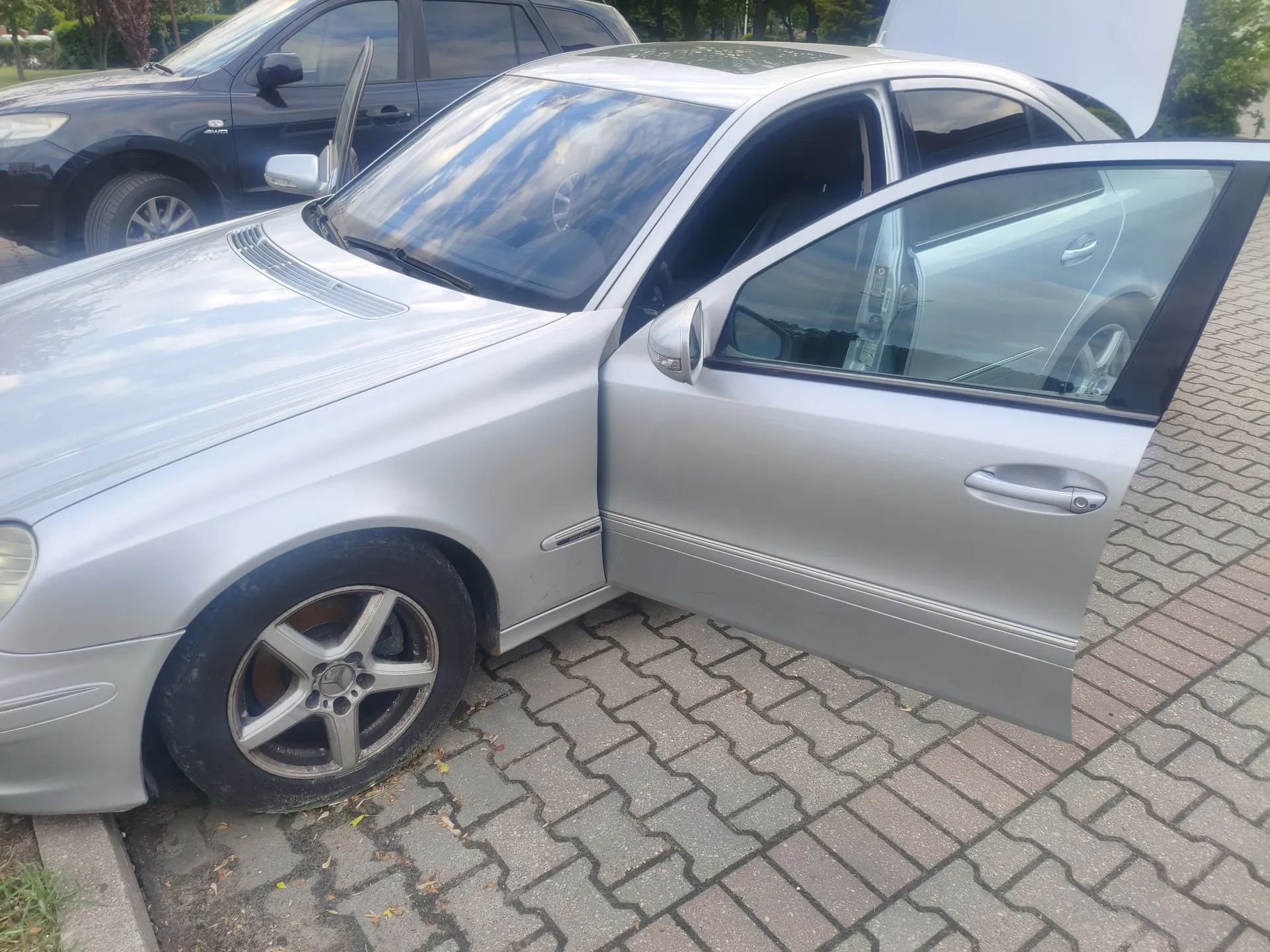 Srebrny samochód osobowy typu sedan z otwartą przednią lewą drzwiami, zaparkowany na brukowanej powierzchni. Widoczne są felgi z emblematem Mercedes-Benz, dach z szybą panoramiczną oraz odbicie nieba w szybach. W tle czarny SUV z otwartymi drzwiami i zielone drzewa. Na nawierzchni leżą rozrzucone liście.
