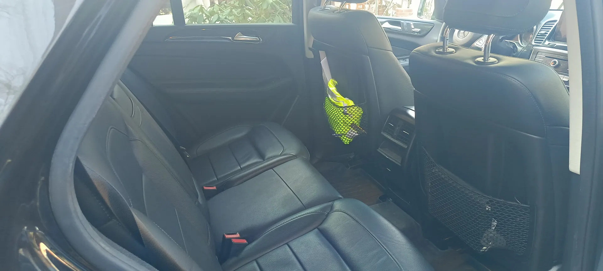 Wnętrze samochodu z czarnymi skórzonymi siedzeniami. Widoczne są tylna część kabiny, otwarte drzwi z uchwytem i oknem, przez które widać zielone rośliny na zewnątrz. Na oparciu przedniego siedzenia zamocowana jest zielona siateczka z żółtym przedmiotem.