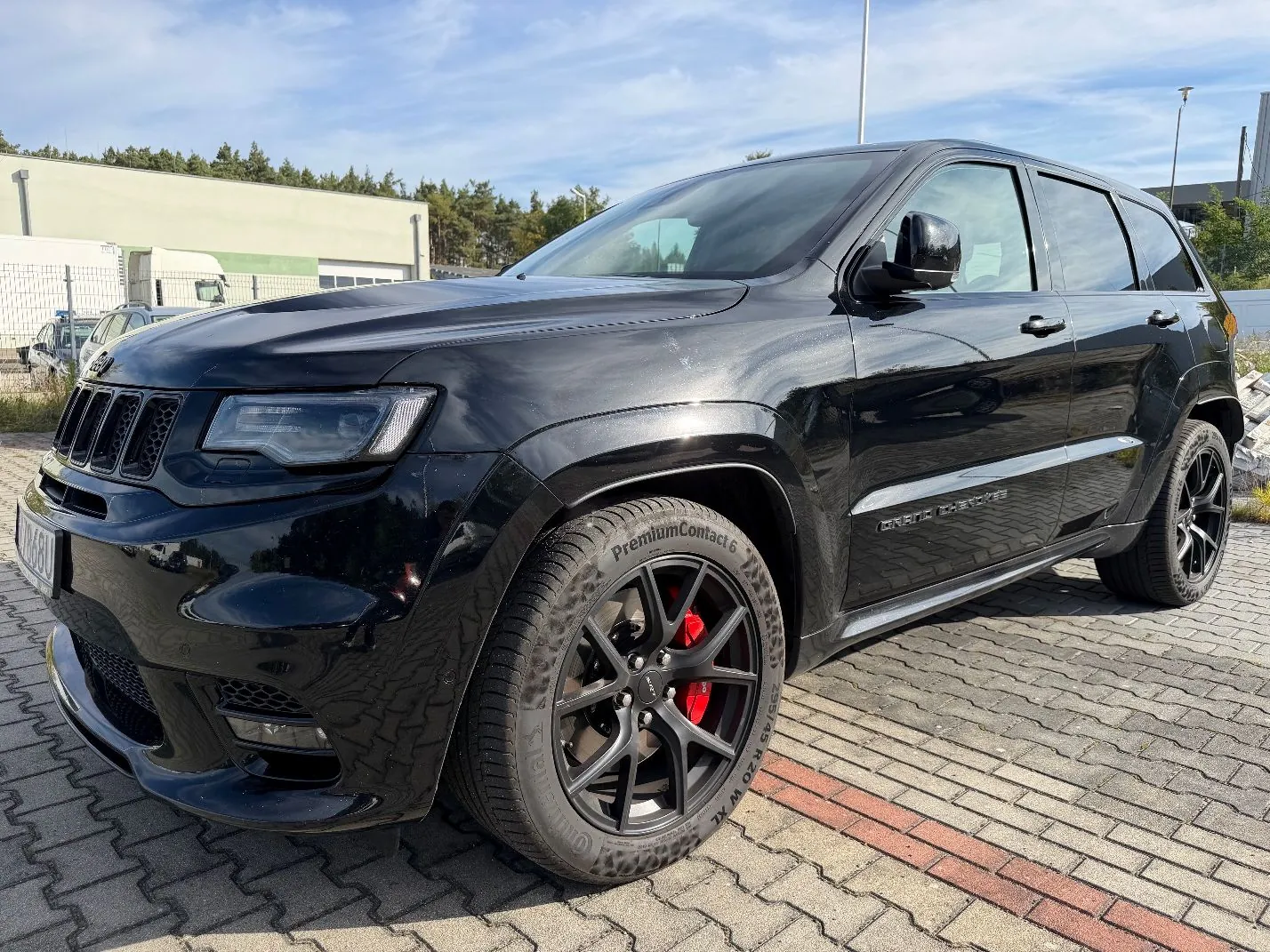 Czarny samochód marki Jeep Grand Cherokee zaparkowany na kostce brukowej. Pojazd jest widoczny z przodu i z boku, posiada czarne felgi z czerwonymi zaciskami hamulcowymi oraz opony Continental PremiumContact 6. W tle widoczny jest jasny budynek przemysłowy oraz niebo z lekkimi chmurami.