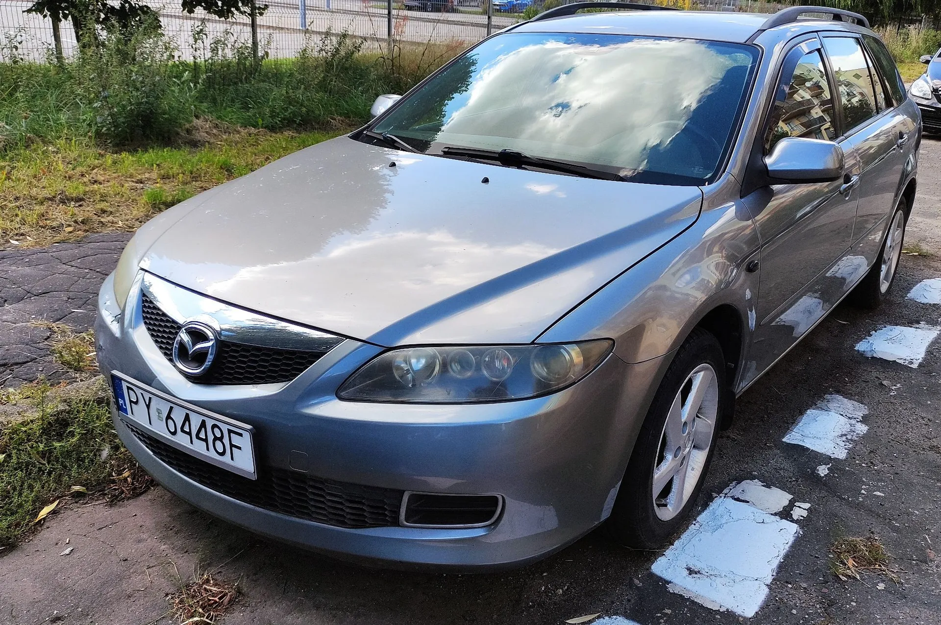 Srebrny samochód marki Mazda w nadwoziu kombi, zaparkowany na utwardzonej nawierzchni obok trawnika. Widoczna jest przednia część pojazdu z tablicą rejestracyjną o numerze PY 6448F. W tle widać zielone krzewy i ogrodzenie.