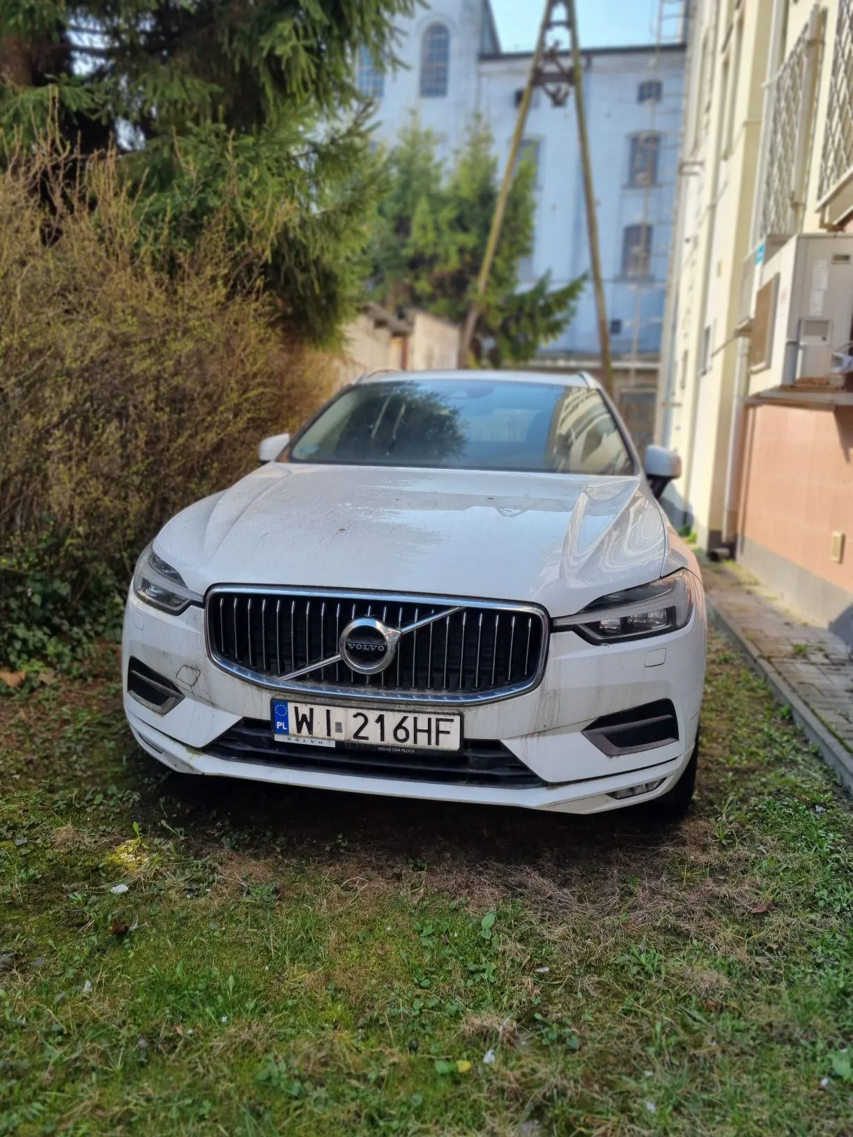Biały samochód marki Volvo z tablicą rejestracyjną WI 216HF zaparkowany na trawie obok budynków. W tle widoczne są drzewa, krzewy oraz budynki mieszkalne z oknami i balkonami. Samochód znajduje się w centralnej części kadru, a tablica rejestracyjna jest czytelna.