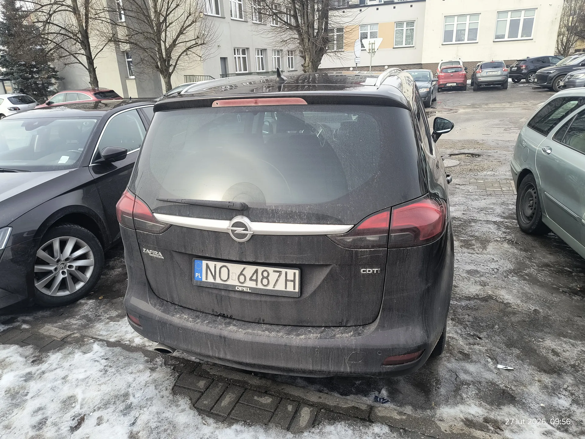 Zdjęcie przedstawia tył czarnego samochodu Opel Zafira z tablicą rejestracyjną PL NO 6487H. Samochód jest zabrudzony, widoczne są napisy ZAFIRA, OPEL oraz CDTI. W tle parking z innymi pojazdami, budynkami mieszkalnymi, drzewami bez liści oraz śniegiem na ziemi. W prawym dolnym rogu widoczny znaczek z datą 27 lut 2026, 09:56.