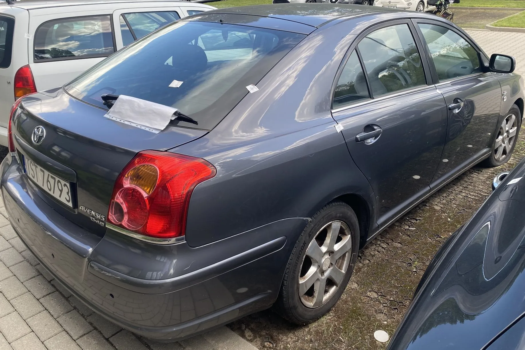 Szary samochód osobowy marki Toyota Avensis zaparkowany na placu z kostki brukowej. Widoczny jest tył i prawy bok pojazdu. Samochód posiada tablicę rejestracyjną o numerze IST 76793 oraz napis AVENSIS D-4D na klapie bagażnika. Wewnątrz auta, na tylnej półce, znajduje się biała kartka papieru. Obok stoją inne samochody.