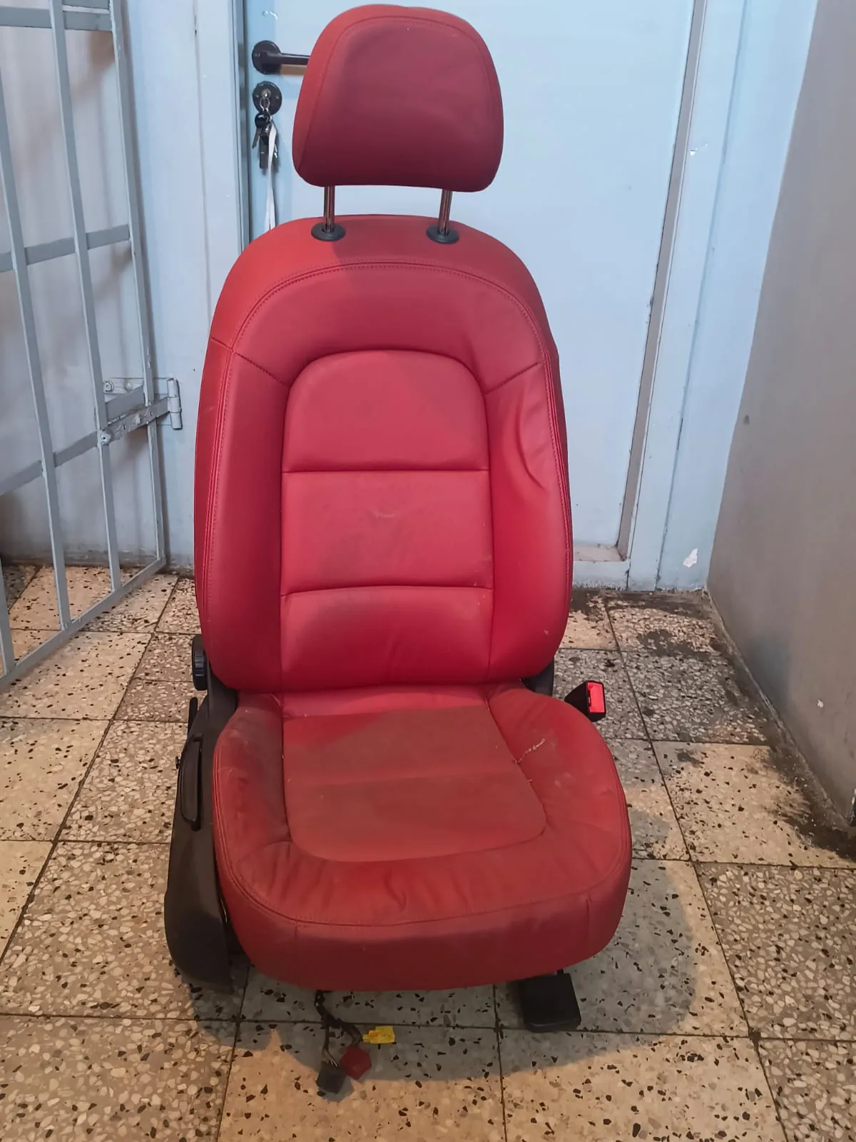 Czerwony, skórzany fotel samochodowy z zagłówkiem, ustawiony pionowo na jasnej, płytkowej podłodze na tle jasnoniebieskich drzwi i metalowej kraty.