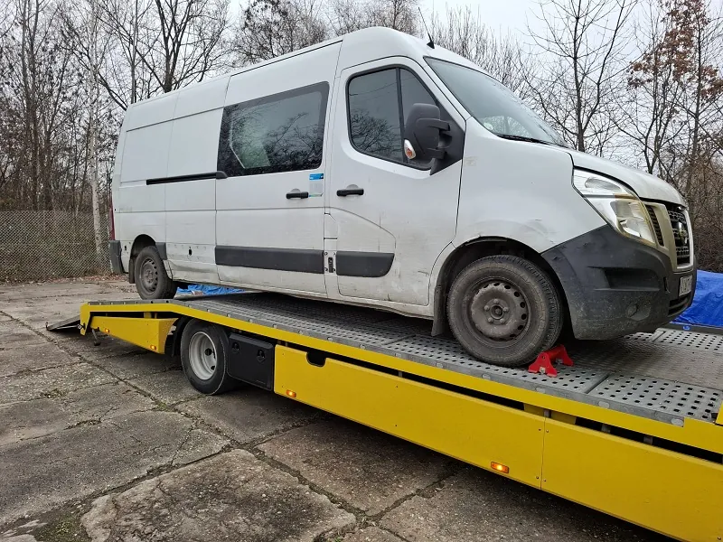 Biały furgon z czarnymi elementami na przednim zderzaku i bokach, umieszczony na żółtej płycie przyczepy holowniczej z metalową siatką. Furgon jest zabezpieczony czerwonymi klinami pod przednimi kołami. Tło stanowią bezlistne drzewa, siatka ogrodzeniowa oraz betonowa powierzchnia z pęknięciami. Na przodzie furgona widoczna jest nieczytelna tablica rejestracyjna.