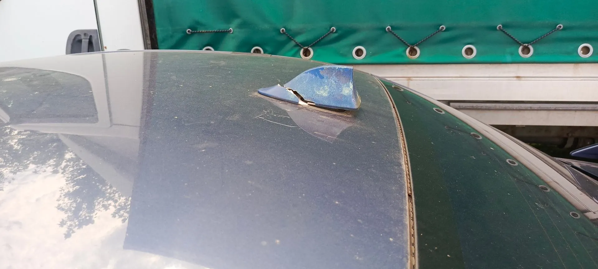 Zbliżenie na dach ciemnoniebieskiego samochodu, na którym znajduje się uszkodzona antena typu płetwa. Plastikowa obudowa anteny jest pęknięta i częściowo oderwana, odsłaniając wnętrze elementu.
