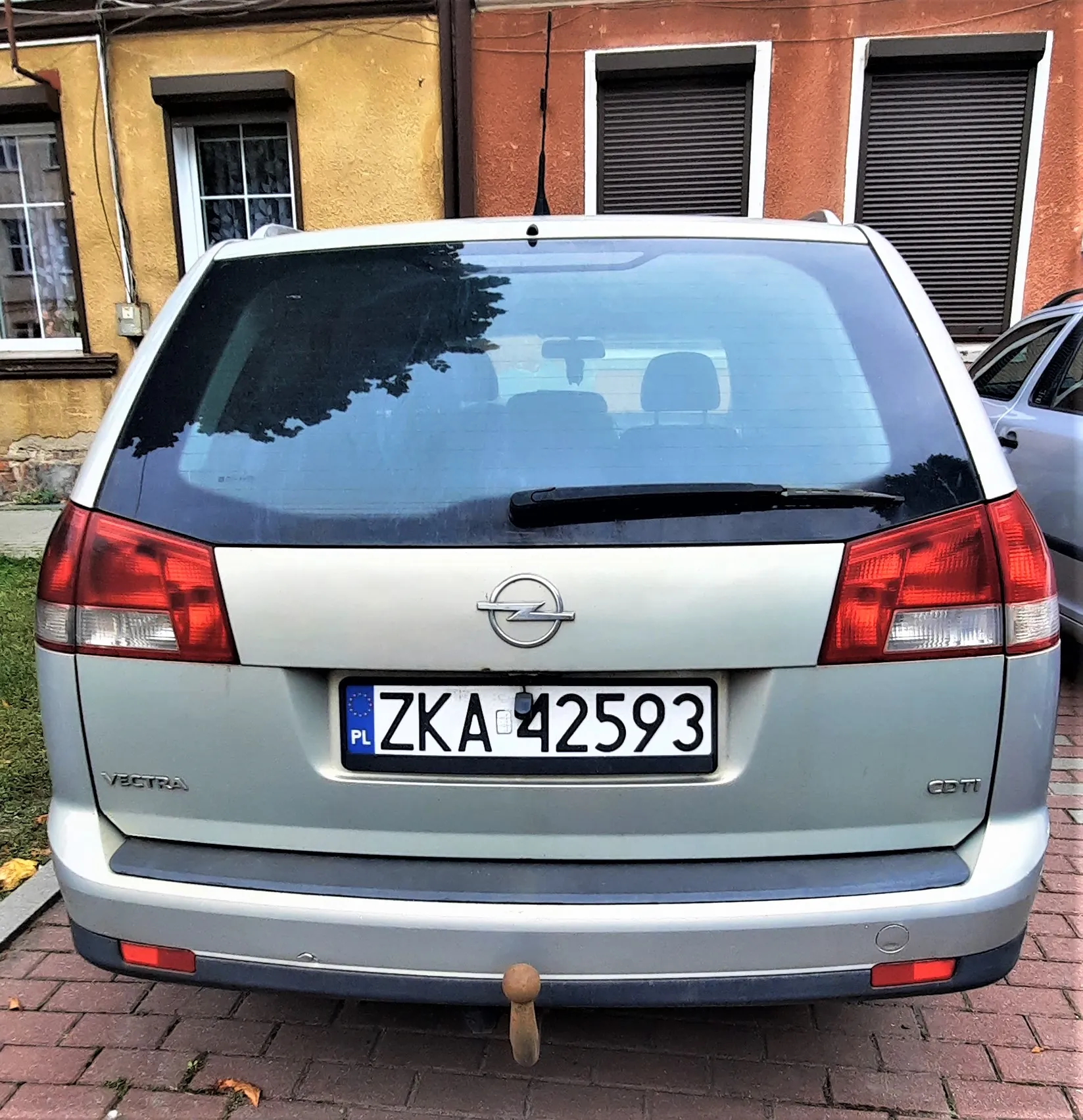 Tylny widok srebrnego samochodu marki Opel Vectra CDTI. Pojazd posiada polską tablicę rejestracyjną o numerze ZKA 42593. Samochód zaparkowany jest na kostce brukowej przed budynkiem o żółtej i pomarańczowej elewacji z oknami i roletami.