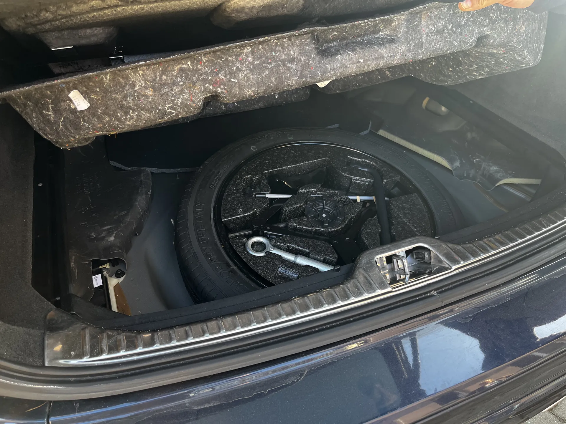 Otwarty bagażnik samochodu z umieszczonym w środku kołem zapasowym. Wokół koła znajdują się narzędzia: klucz, podnośnik i czarne piankowe elementy mocujące. Wewnętrzna część pokrywy bagażnika wyłożona jest szarą pianką z widoczną białą etykietą. Zewnętrzna część bagażnika ma czarny lakier z metalową listwą. Na lewej stronie widoczny jest fragment mechanizmu z białą etykietą.