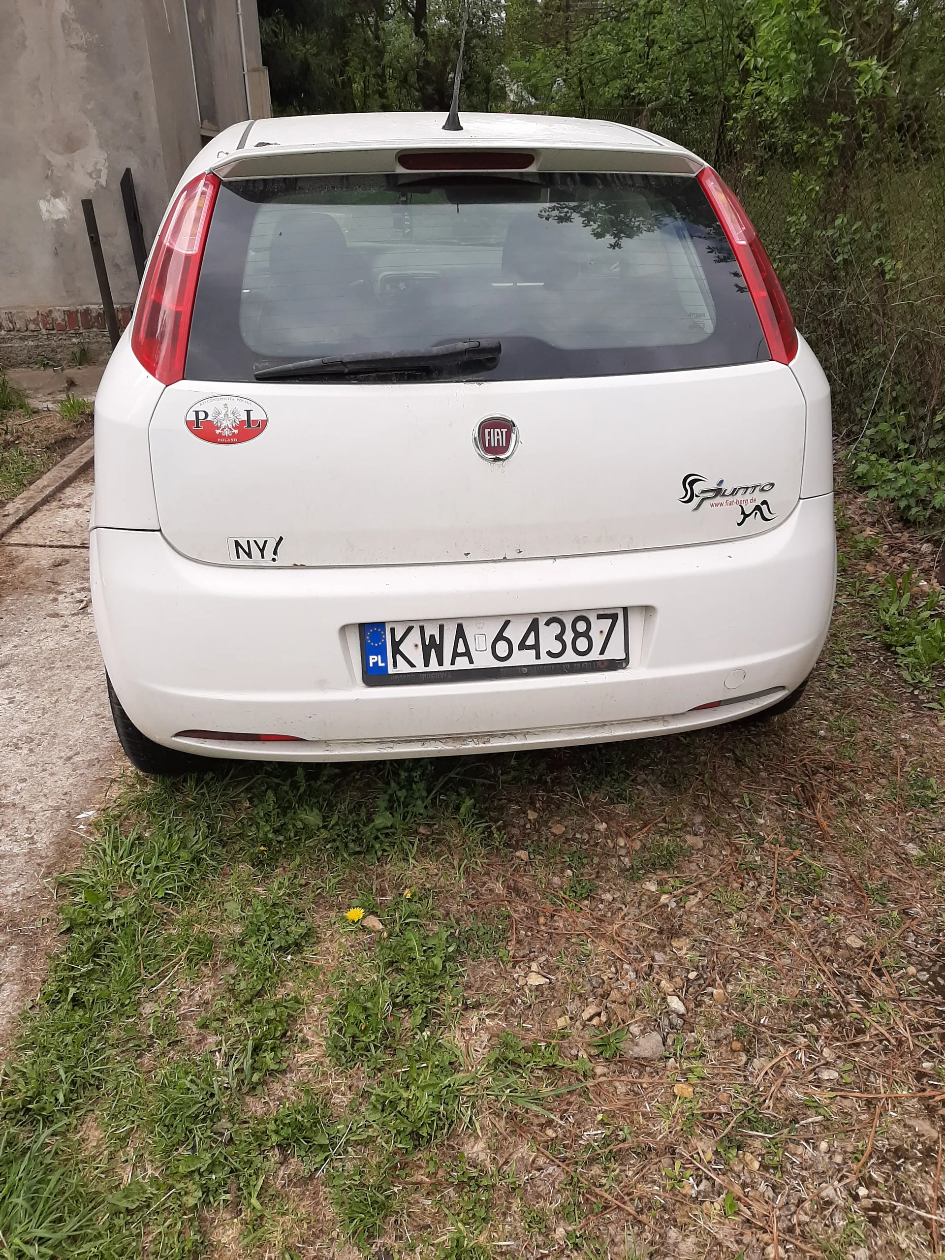 Biały samochód marki Fiat z tyłu, zaparkowany na drodze zagrzewanej. Widoczna tablica rejestracyjna KWA 64387 z niebieskim pasem PL po lewej stronie. Na bagażniku znajdują się naklejki: czerwona z napisem PL i herbem, czarna z napisem NYI oraz czarne logo z napisem Sunro. W tle zielona trawa, drzewa i fragment ogrodzenia. Samochód jest nieco zabrudzony.
