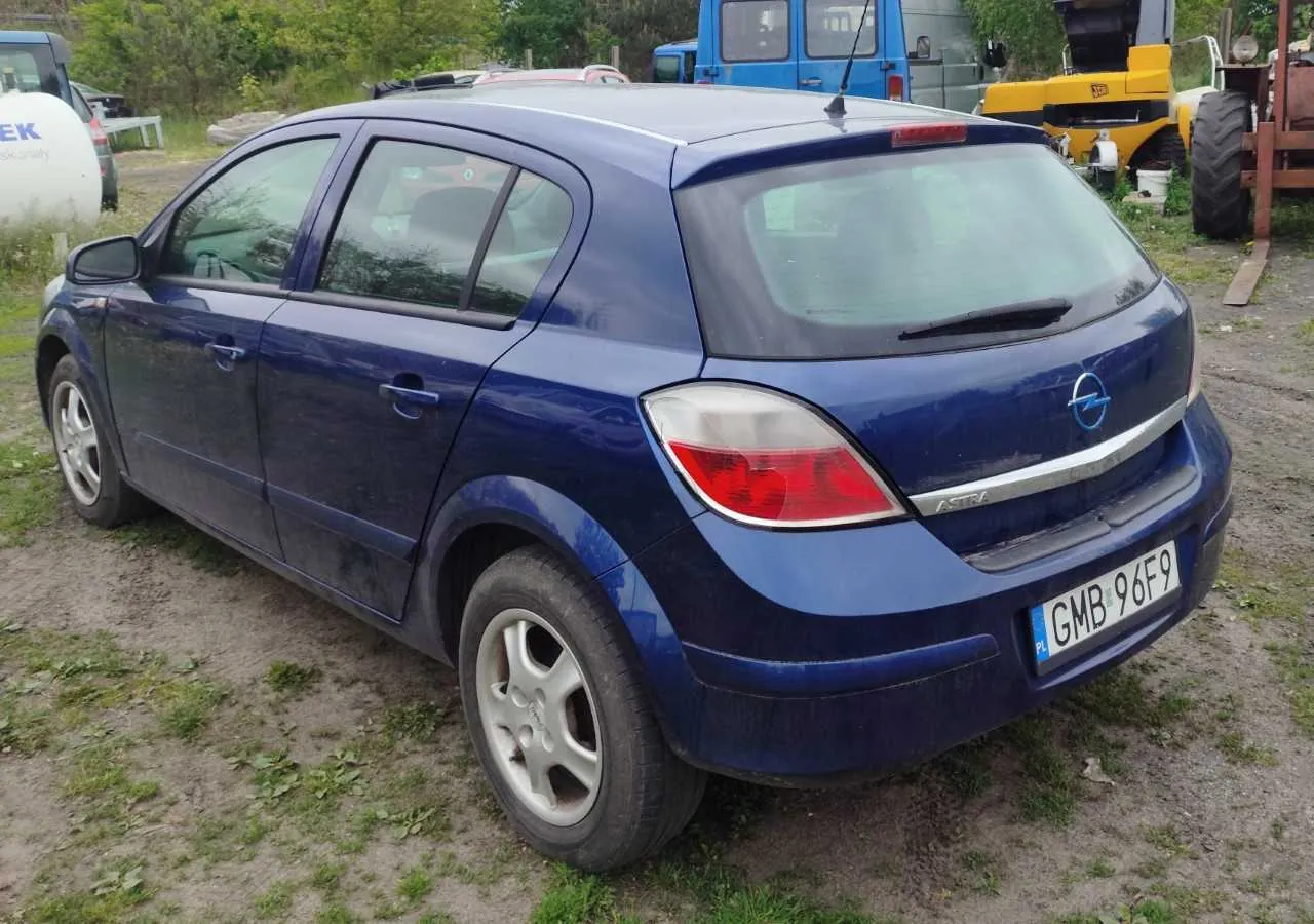 Niebieski samochód Opel Astra hatchback z tyłu, z widoczną tablicą rejestracyjną PL GMB 96F9. Pojazd stoi na podwórku z drobnej ziemi i trawy. W tle widoczne są inne pojazdy: niebieski ciężarówka, żółty traktor oraz biały zbiornik z częściowo widocznym napisem EK. Samochód ma srebrne felgi i czerwone światła tylnego. Na tylnej szybie umieszczono logo Opel oraz napis ASTRA.