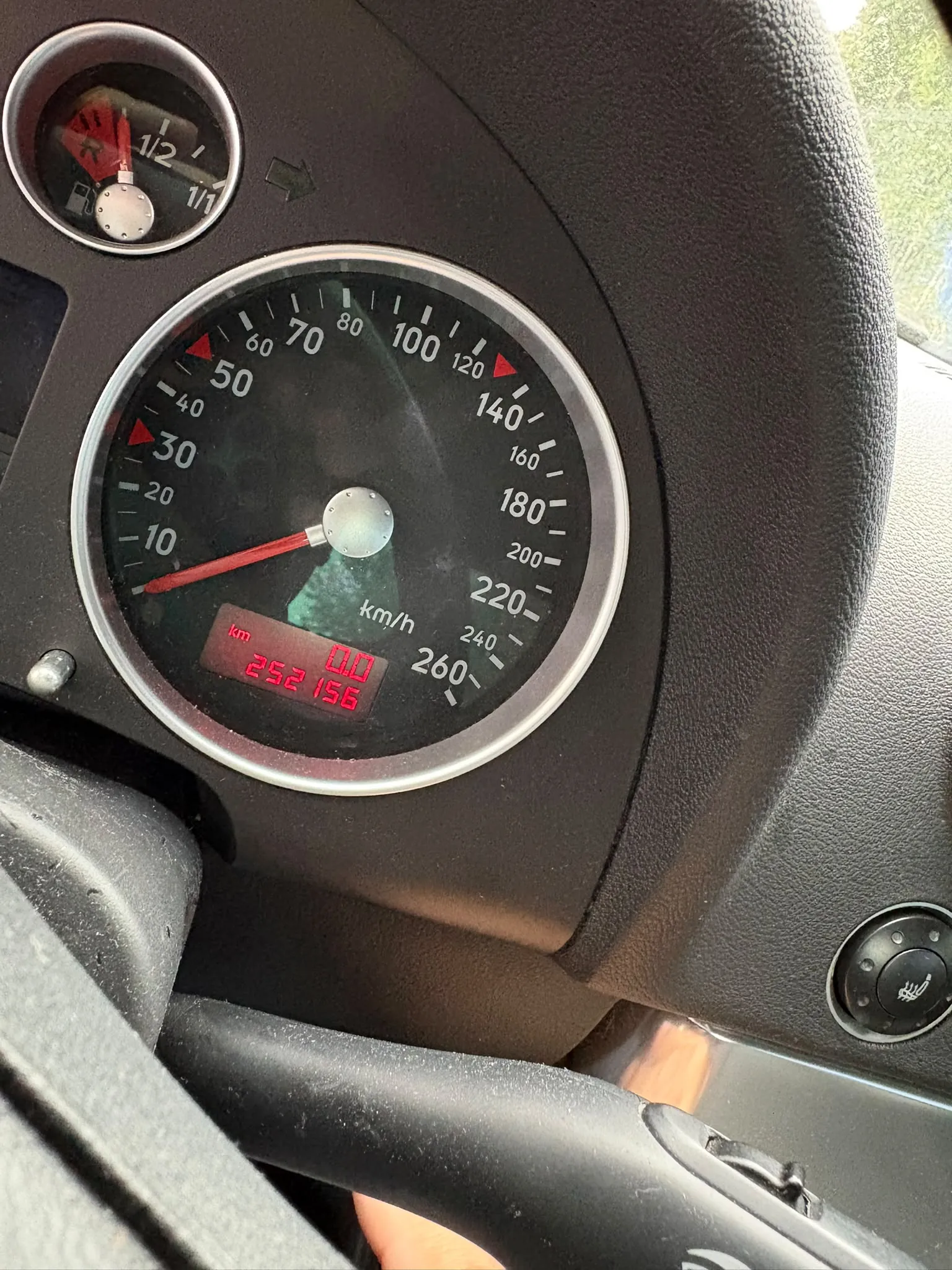 Zbliżenie na deskę rozdzielczą samochodu. Głównym elementem jest okrągły prędkościomierz z tarczą wyskalowaną do 260 km/h, z wskazówką wskazującą wartość bliską 0. Poniżej tarczy znajduje się cyfrowy licznik przebiegu wyświetlający czerwoną liczbę 252156. W lewym górnym rogu widoczny jest fragment wskaźnika poziomu paliwa. Całość osadzona jest w ciemnoszarej, plastikowej obudowie kokpitu.