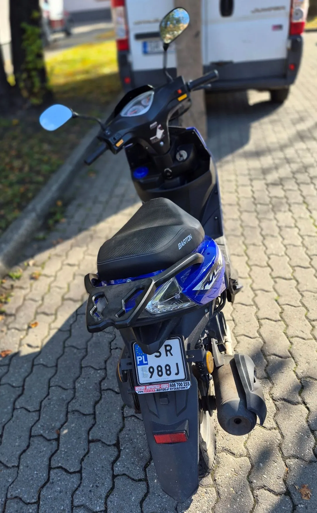 Niebieski skuter marki Barton zaparkowany na kostce brukowej. Widok z tyłu pojazdu, ukazujący siedzenie, tylną lampę oraz tablicę rejestracyjną o numerze SR 988J z oznaczeniem PL. W tle widoczny jest biały samochód dostawczy.