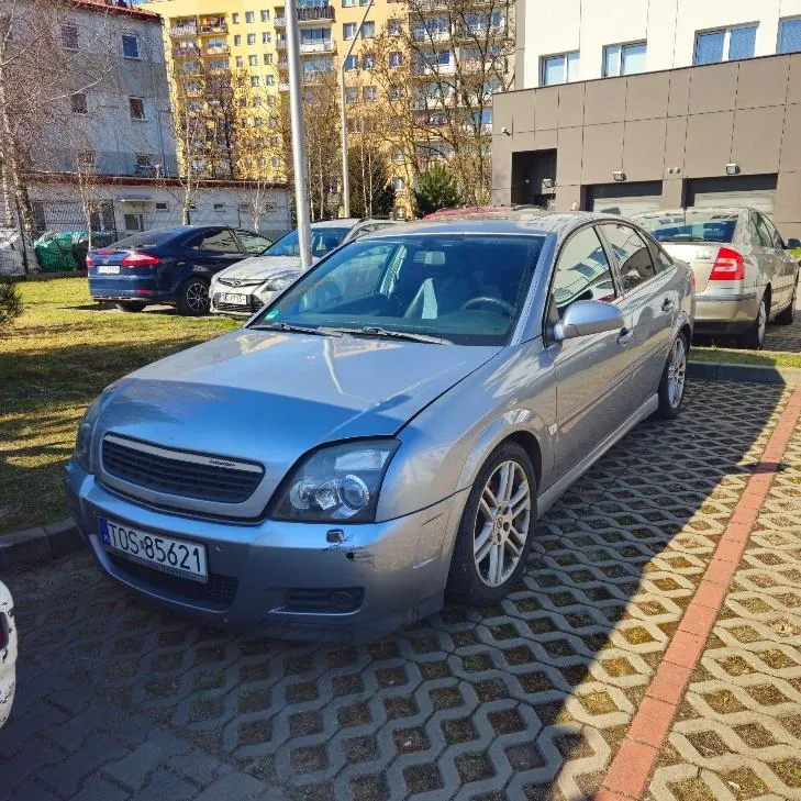 Srebrny samochód osobowy marki Opel (model Vectra) zaparkowany na podwórku z kostki brukowej. Widoczna tablica rejestracyjna TOS-85621. W tle inne zaparkowane samochody, budynki mieszkalne (w tym żółty blok wielorodzinny), drzewa bez liści oraz budynek z garażami. Słoneczna pogoda, cień pada na jezdnię.