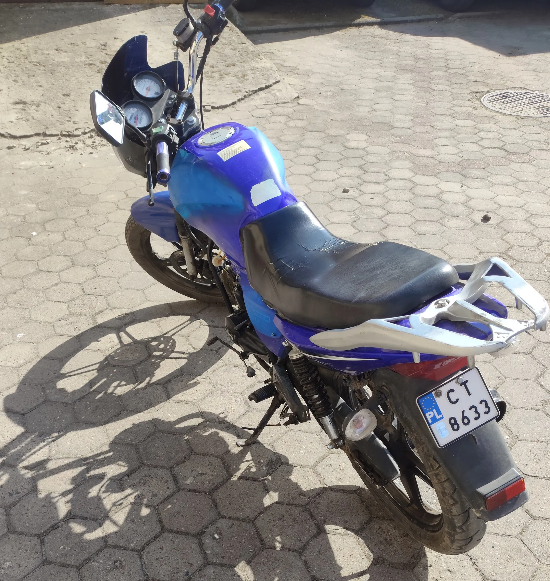 Niebieski motocykl zaparkowany na brukowanej powierzchni z sześciokątnymi płytami. Widoczna tablica rejestracyjna z napisem 'PL CT 8633'. Motocykl posiada czarne siedzenie, dwa okrągłe wskaźniki na kierownicy oraz lusterko wsteczne po lewej stronie. W tle znajduje się kanałowa pokrywa i betonowa powierzchnia.