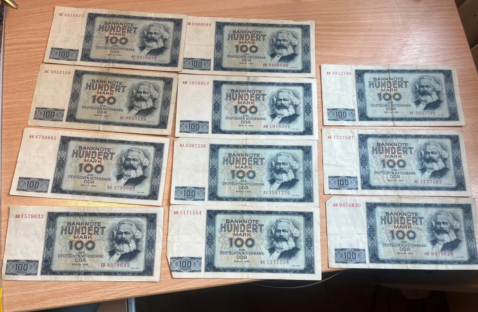 Kolekcja dwunastu banknotów o nominale 100 marek z Niemieckiej Republiki Demokratycznej (DDR), ułożonych w trzech rzędach po cztery sztuki na drewnianym blacie. Każdy banknot ma niebiesko-szary kolor, zawiera portret mężczyzny z brodą, napis 'BANKNOTE HUNDERT MARK 100' oraz tekst 'DEUTSCHEN NOTENBANK DDR'. Na banknotach widoczne są różne numery seryjne.