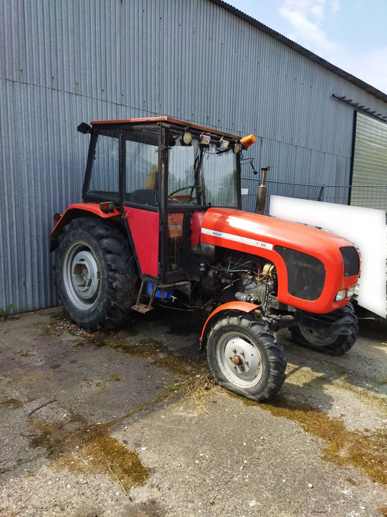 Czerwony traktor marki Ursus, model C-360, z zamkniętą kabiną, zaparkowany na betonowym podłożu przed szarym, falistym budynkiem gospodarczym. Na maszynie widoczne są napisy 'URSUS' oraz 'C-360'.