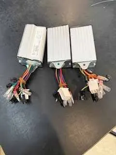 Trzy prostokątne urządzenia elektroniczne o srebrnym kolorze ustawione na ciemnej powierzchni. Każdy z nich posiada przewody w różnych kolorach (czerwony, żółty, czarny, biały) podłączone do złącz. Przewody zwisają swobodnie w dół. Tło stanowi ciemna, jednolita powierzchnia.