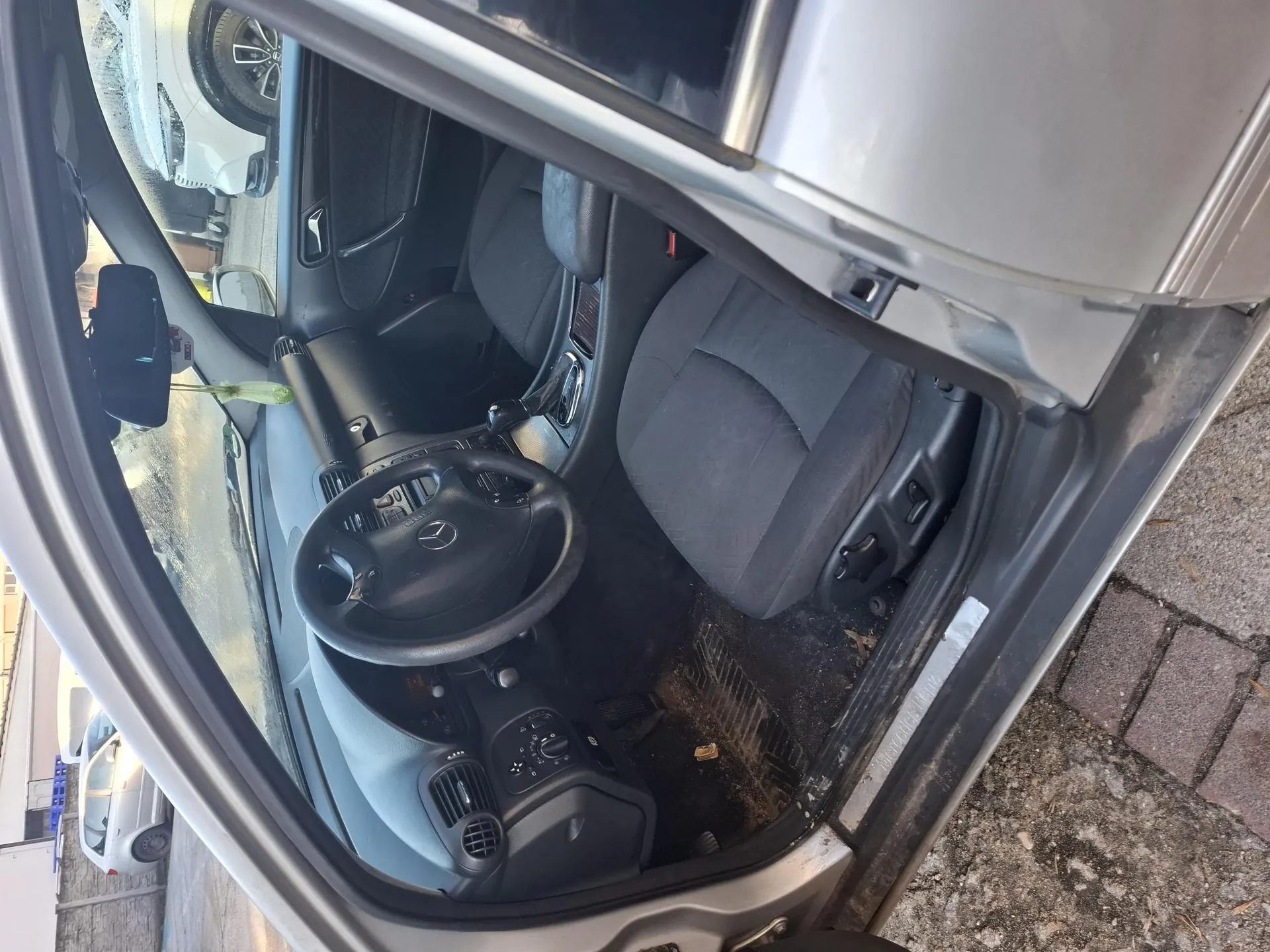 Zdjęcie przedstawia wnętrze samochodu marki Mercedes z otwartą przodem drzwiami. Widoczne są czarne siedzenia, kierownica z logo Mercedes-Benz, czarna deska rozdzielcza z przyciskami i wskaźnikami. Na podłodze wokół pedałów znajduje się brud i liście. W tle za przednią szybą widoczne są zaparkowane samochody: srebrny i biały SUV. Na lusterku wstecznym wiszą zielony aromatyzator. Zewnętrzna powierzchnia pod samochodem to brukowana droga.
