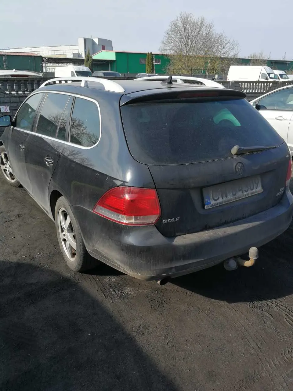 Ciemnoszary samochód osobowy typu kombi marki Volkswagen Golf, widok od tyłu. Pojazd jest zabrudzony, posiada białe dachowe pręty i czerwone światła tylnego. Na tablicy rejestracyjnej widoczny napis PL oraz numer C11 04309. W tle stoją inne pojazdy, zielony budynek, drzewa bez liści i ogrodzenie. Asfaltowa powierzchnia parkingu z widocznymi odciskami opon.