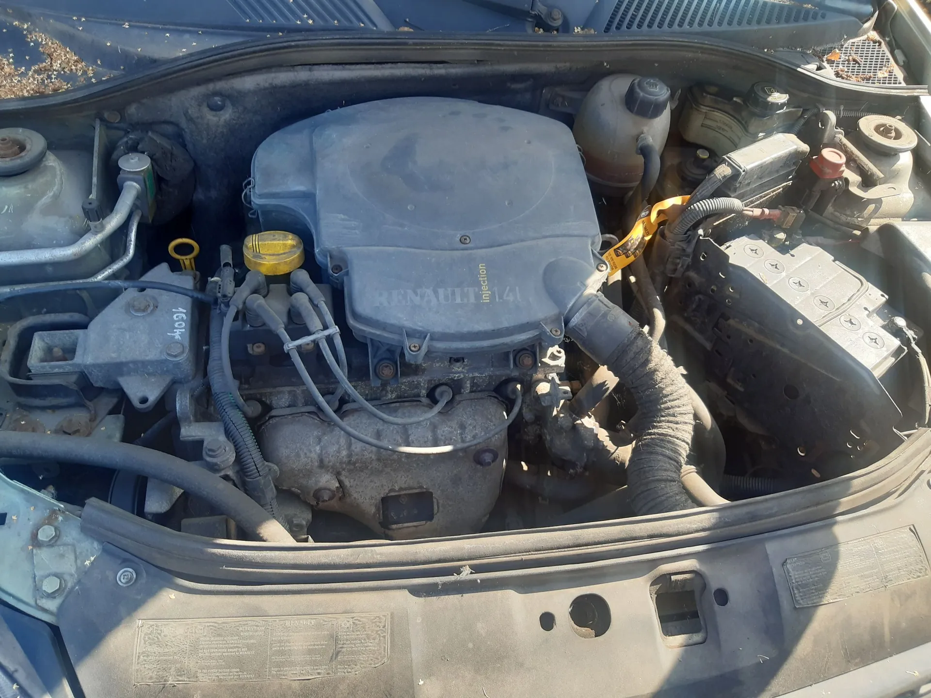 Widok z góry na komorę silnika samochodu marki Renault. W centrum znajduje się szara osłona silnika z napisem 'RENAULT injection 1.4'. Po prawej stronie widoczny jest akumulator oraz zbiornik z płynem. Całość jest zakurzona.