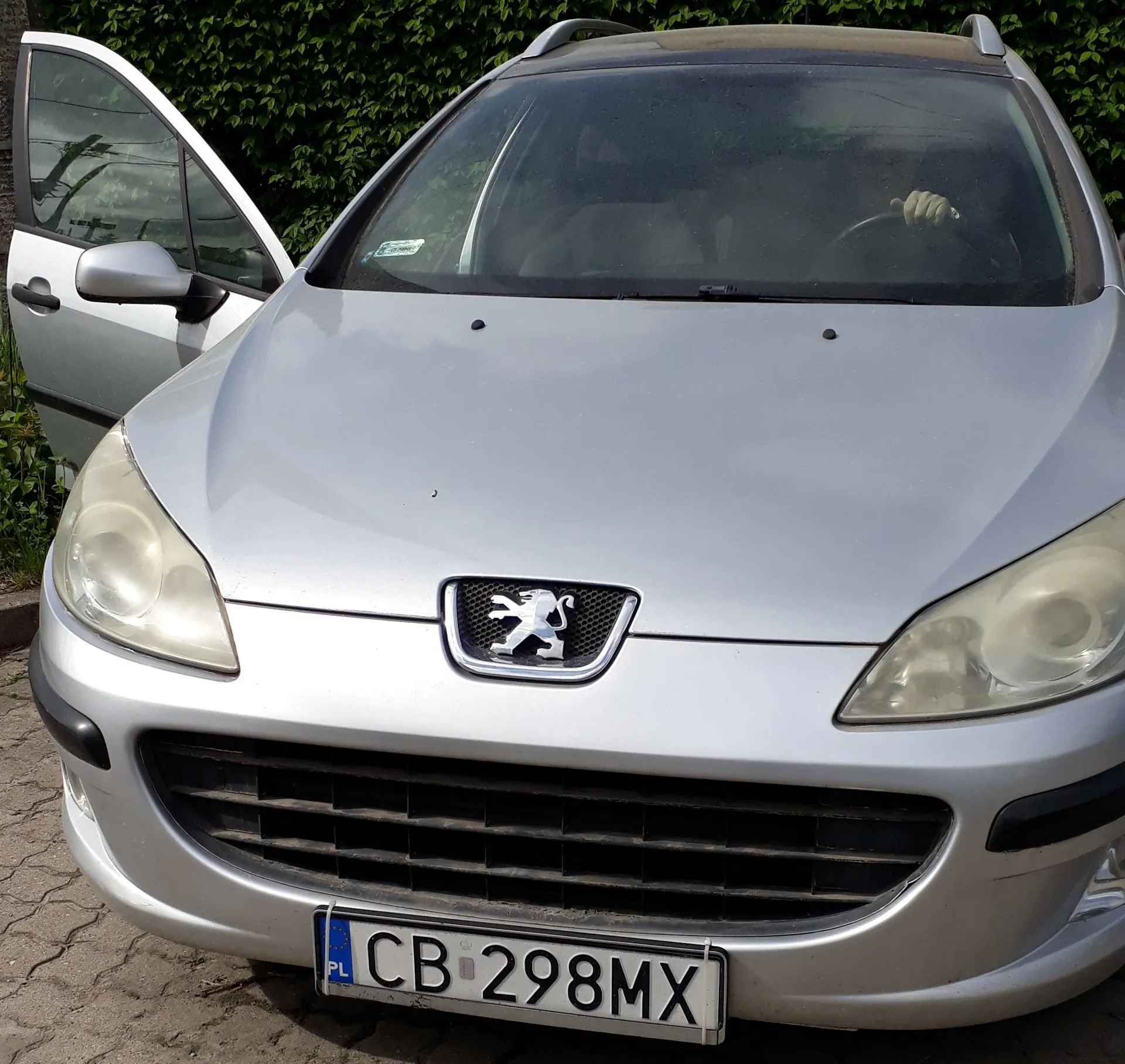 Srebrny samochód marki Peugeot zaparkowany przodem do widza. Widoczny jest przód pojazdu z logo lwa na masce oraz tablica rejestracyjna o numerze CB 298MX. Lewe przednie drzwi samochodu są otwarte. W tle znajduje się zielona ściana z roślinności.