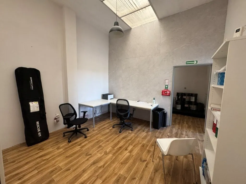 Jasne pomieszczenie biurowe z drewnianą podłogą. W centrum znajdują się dwa białe biurka ustawione w kształcie litery L, przy których stoją dwa czarne krzesła biurowe na kółkach. Na biurkach znajduje się drukarka oraz drobne przedmioty. Po lewej stronie stoi czarny pokrowiec z napisem „SIEMENS Games” oraz „RESCUE KIT”. Po prawej stronie widoczny jest biały regał z dokumentami oraz białe krzesło. W tle znajduje się otwarte przejście do innego pomieszczenia, nad którym wisi zielona tabliczka ewakuacyjna ze strzałką w prawo. Na ścianie obok przejścia zamontowany jest czerwony przycisk alarmowy. Z sufitu zwisa jedna szara lampa.**