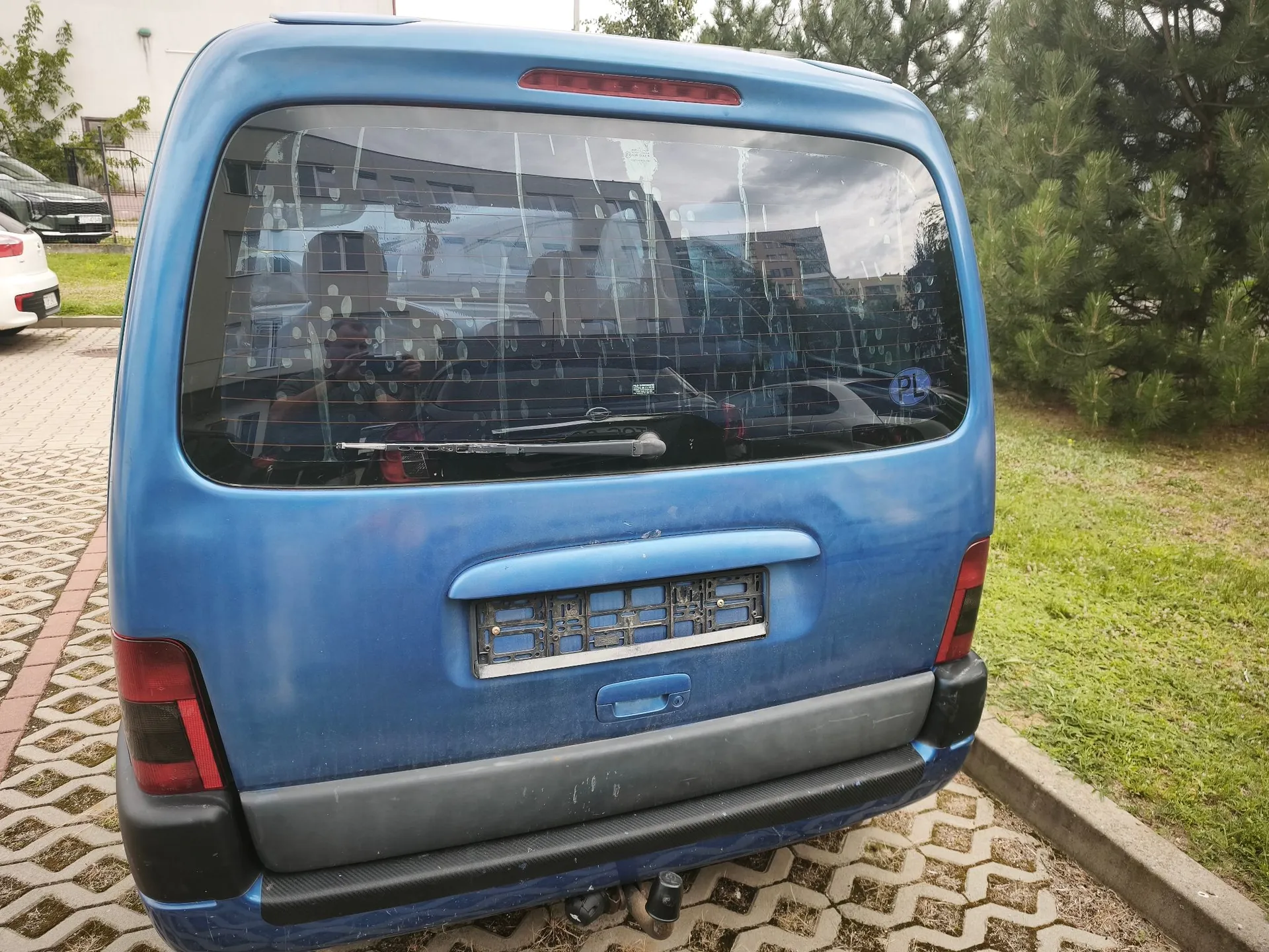 Tylny widok niebieskiego samochodu typu van zaparkowanego na kostce brukowej. Pojazd jest brudny, a tylna szyba posiada liczne zacieki. Samochód nie posiada tablicy rejestracyjnej, widoczny jest jedynie pusty uchwyt na tablicę. Po prawej stronie znajduje się trawnik i zielone drzewa.