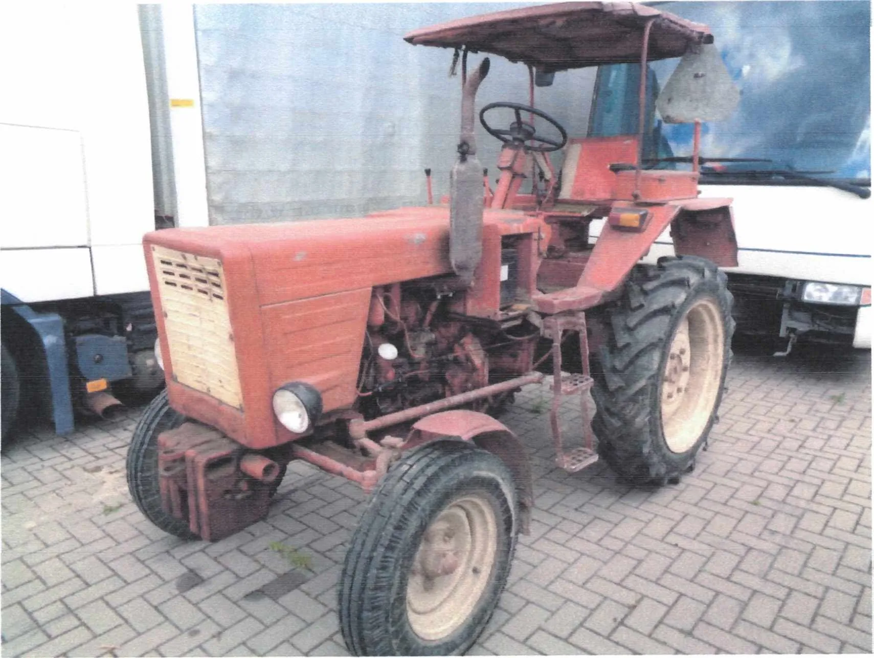 Czerwony traktor o widocznych oznakach zużycia, z daszkiem chroniącym przed słońcem, kierownicą, dużymi oponami z tyłu i mniejszymi z przodu. Na przedniej części traktora znajduje się reflektor oraz siatkowana osłona chłodnicy. W tle widoczne są częściowo zasłonięte pojazdy: biały ciężarówka po lewej stronie i biały autobus po prawej. Podłoże stanowi brukowana powierzchnia.