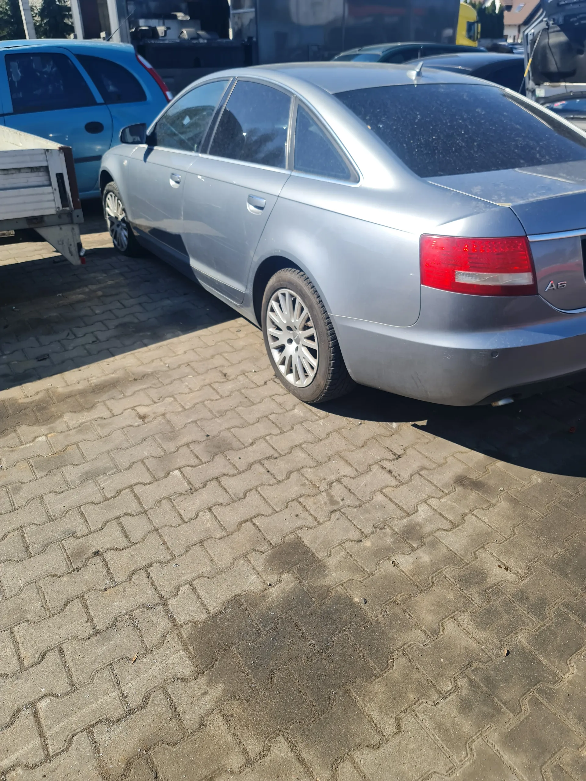 Srebrny samochód Audi A6 zaparkowany na brukowanej powierzchni. Widoczne są czerwone światło tylnego reflektora, srebrne felgi oraz napis 'A6' na bagażniku. Obok znajduje się niebieski samochód. W tle budynki i fragmenty innych pojazdów. Brukowana powierzchnia składa się z kamiennych płyt ułożonych w wzór herringbone.