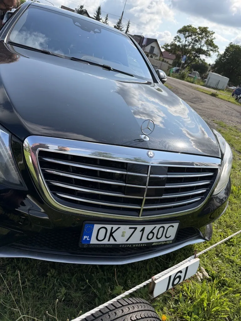 Zbliżenie na przód czarnego samochodu marki Mercedes-Benz. Widoczna jest maska z logo marki, chromowany grill oraz tablica rejestracyjna o numerze OK 7160C z oznaczeniem PL. Samochód stoi na trawie obok białej linii odgradzającej teren, na której znajduje się tabliczka z numerem 161.