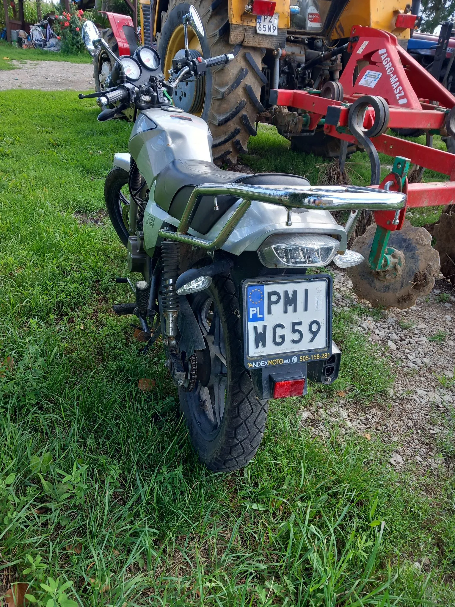 Zdjęcie przedstawiające tył srebrnego motocykla zaparkowanego na trawie obok czerwonej maszyny rolniczej. Motocykl posiada tablicę rejestracyjną o numerze PMI WG59 z oznaczeniem PL. Poniżej tablicy widnieje napis 'ANDEXMOTO.eu' oraz numer telefonu '505-158-281'. W tle widoczna jest część traktora z żółtą tablicą rejestracyjną PZ 85N9 oraz czerwona maszyna marki AGRO-MASZ.