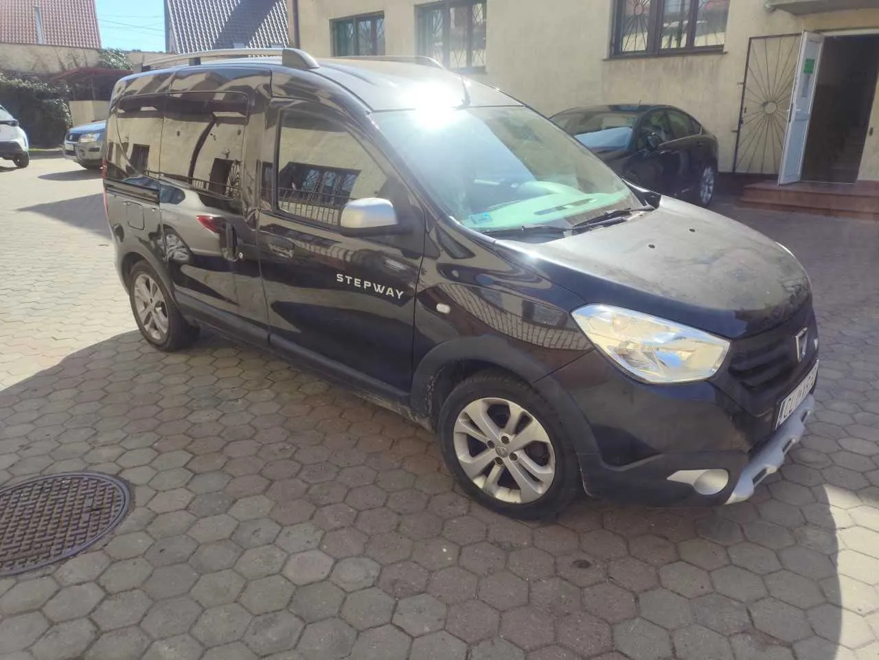 Czarny samochód typu van z napisem STEPWAY na boku, zaparkowany na brukowanej powierzchni z sześciokątnymi płytami. W tle widoczne są budynki z oknami i drzwiami, inne pojazdy oraz słońce oświetlające przednią szybę. Na lewo od samochodu znajduje się okrągła studzienka kanalizacyjna.