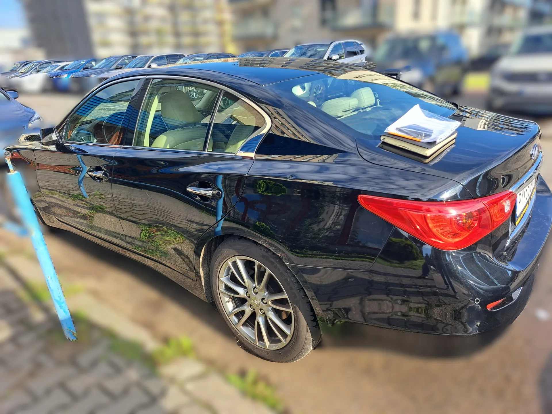 Czarny samochód osobowy typu sedan zaparkowany na zewnątrz. Widok z boku i z tyłu. Na tylnej półce samochodu leżą dokumenty i folder. W tle widoczne są inne zaparkowane samochody oraz budynki mieszkalne.
