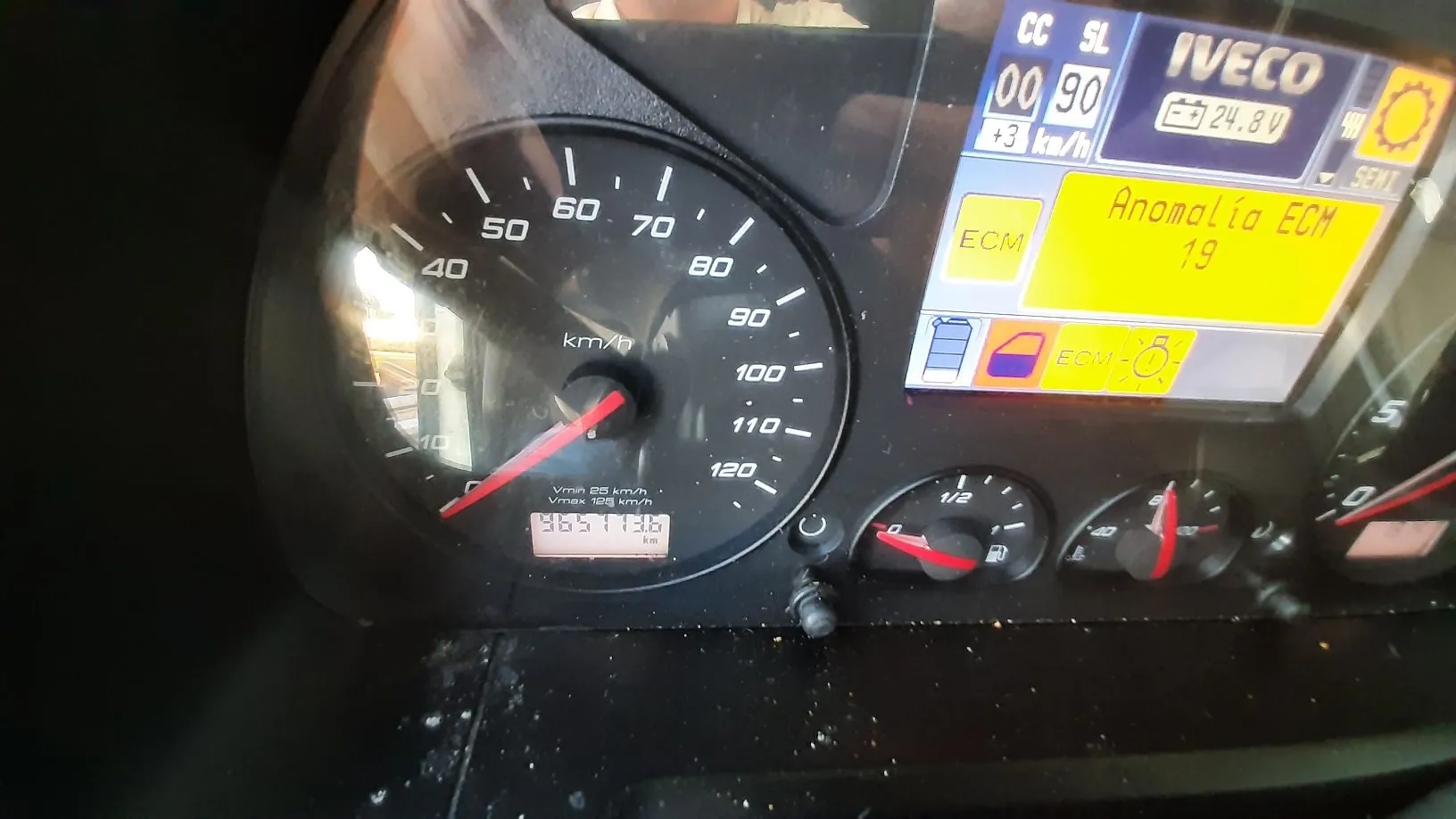 Zbliżenie na deskę rozdzielczą pojazdu marki Iveco. Widoczny jest prędkościomierz z maksymalną skalą 120 km/h, wskazówka wskazuje wartość bliską 0. Poniżej znajduje się licznik kilometrów z wartością 965113,6 km. Po prawej stronie znajduje się wyświetlacz cyfrowy z napisem 'IVECO', napięciem akumulatora '24.8V' oraz żółtym komunikatem ostrzegawczym 'Anomalía ECM 19'. Widoczne są również mniejsze wskaźniki poziomu paliwa i temperatury.